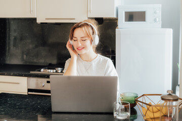 Dreaming young Woman in headset with closed eyes enjoying and listen to music while working on laptop at Kitchen Table in tiny flat. Take a break for recovery, mental health. Relax, no stress concept