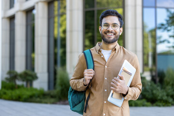 Smiling hispanic student stands outside academic building holding backpack and notebooks. Concept...