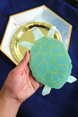 Hand Holding Green Blue Gold Turtle Napkin For Under The Sea Ocean Theme Unisex Boys Girls Kids Childrens Birthday Party Set Over Table