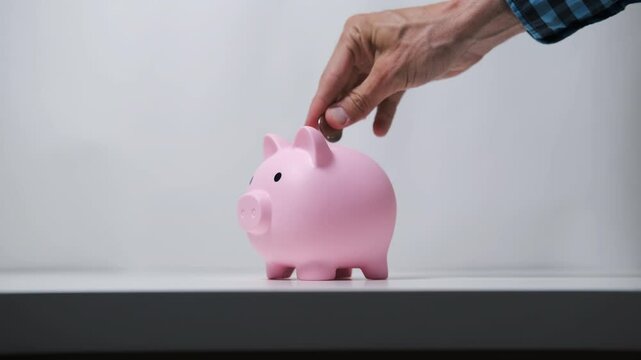 Male hand inserts coins into a pink piggy bank against a white backdrop, emphasizing savings and financial stability
