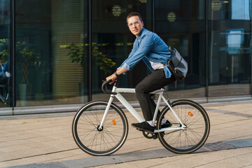 Fototapeta premium A young man riding a bicycle past a modern office building, embodying an active and healthy urban lifestyle.