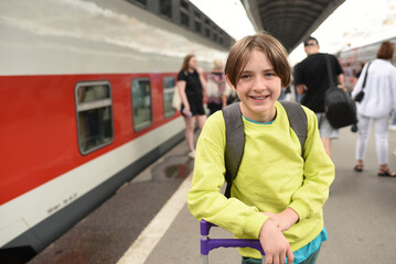 happy 11 year old boy with a backpack and suitcase on the platform, station stands near a double-decker train.