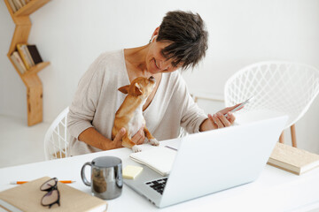 Cheerful woman holding small dog and smartphone near laptop at home