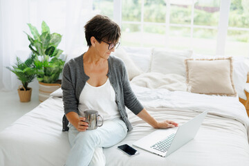 Mature freelancer holding coffee cup and using laptop on bed at home