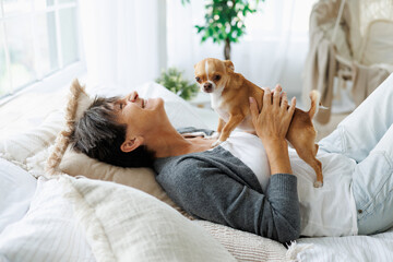 Happy mature woman holding cute dog while relaxing on bed