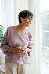 Portrait of relaxed mature woman holding glass of water near window at home