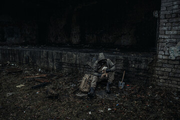 A man in dark green camouflage and a gas mask sits among the ruins of the city. The concept of survival after doomsday or in wartime. Tired lonely soldier.
