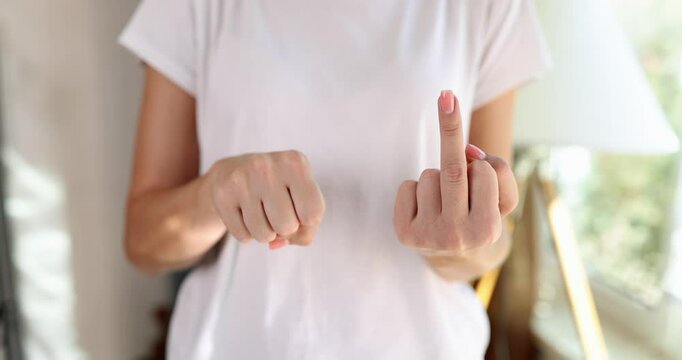 Woman in white t-shirt shows middle finger to camera standing near bright window. Female person makes obscene hand gesture slow motion