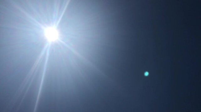 Brilliant Sun in Clear Blue Sky with Lens Flare and Orb Reflection - Natural Light Phenomenon in Midday Summer Sky - Radiant Sunshine Beams and Atmospheric Effects