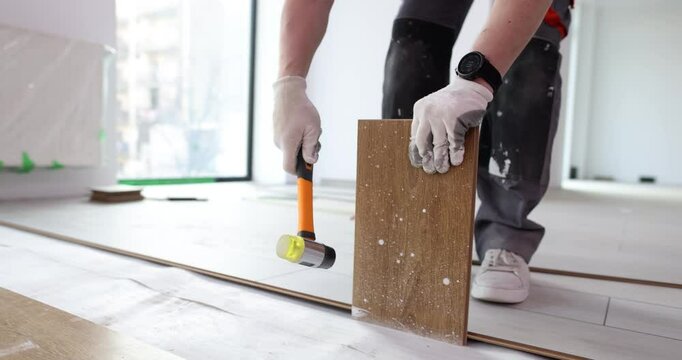 Builder fixes laminate boards tapping details with hammer on floor at construction site. Man installs ground covering in room. Finishing works