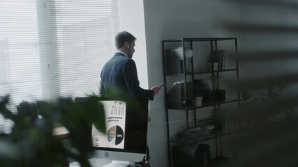Businessman in wireless earbuds walking near window in modern office, holding smartphone and gesturing while talking. Computer monitor showing graphs and charts in foreground