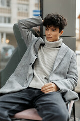 Fashionable young man in grey sweater, white turtleneck, jeans, and black boots sitting on a bench