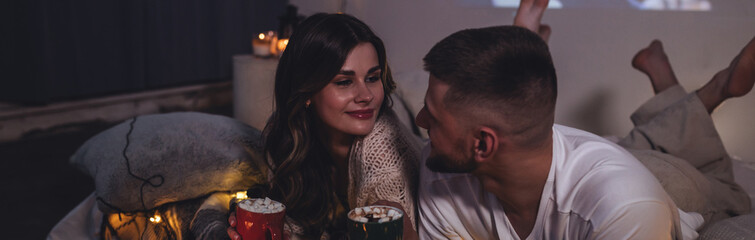 Fototapeta premium Banner. Romantic surprise for girlfriend or boyfriend for Saint Valentine's Day. Bedroom prepared for watching movies decorated with lights, candles. Cozy home Christmas atmosphere, hot chocolate