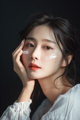 Fototapeta premium Young Asian woman applies moisturizing cream on face for skin hydration in studio setting. Beauty routine captures skincare practice, charming female uses cosmetic product for daily care.