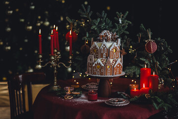 Elegant luxury Christmas table setting for family home party with homemade big gingerbread house, holiday celebration. Red tablecloth, candles, chandelier. Romantic candlelit dinner