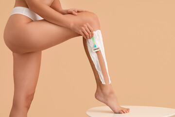 Beautiful young woman with razor and foam shaving legs on beige background, closeup