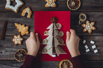 Idea for easy Christmas do it yourself crafts for kids to make. Child's hands with handmade postcards with fir tree and toy from recycled cardboard paper, thread, buttons. Wooden background, flat lay