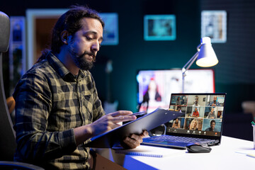 Young guy reviewing his notes while talking with diverse people on a video call, working from home. Serious male individual checks his clipboard while in a virtual meeting on his laptop.