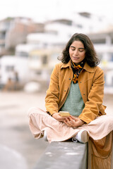 Young woman using smartphone sitting on a wall by the beach