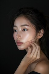 Naklejka premium Asian model woman face close-up beauty studio promo. Applies foundation with finger elegance updo hairstyle. Gaze directed off to side lost in thought observing outside frame.