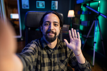 Male influencer waving his hand towards camera, making a daily vlog at home. Smiling young man having video call with friends using modern technology.