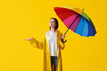 Young happy woman in raincoat with opened colorful umbrella on yellow background