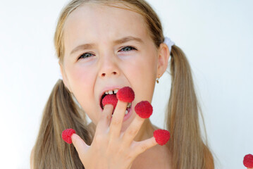 a little cheerful girl with long hair eats red wild raspberries from her fingers