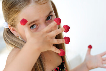 a little cheerful girl with long hair eats red wild raspberries from her fingers