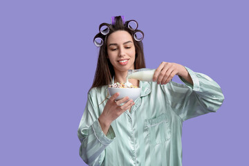 Young woman in pajamas and hair curlers holding milk and cereals on lilac background