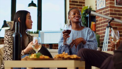 Diverse friends at house party paying attention to interesting story shared by woman. African american guest making asian person laugh in living room while enjoying drinks and charcuterie platter