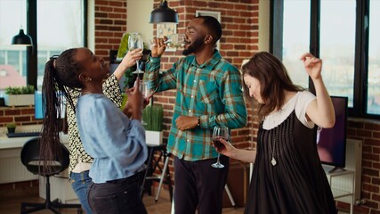 Energetic group of friends dancing at apartment party, having fun together while drinking wine. Multiethnic people doing dance moves on lively music in brick wall living room