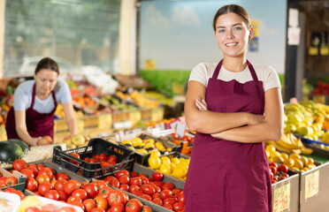 Girl in uniform standing among shelves with fruits and vegetables and looking in camera