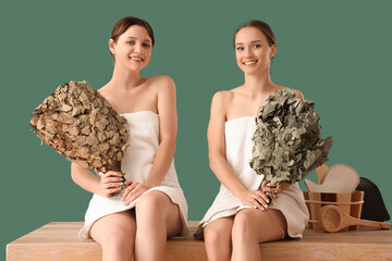 Young women in towels with whisks sitting on bench against green background