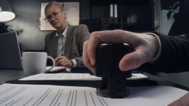 Macro shot of hands of unrecognizable businessman taking document from senior female colleague and putting stamp on it at office desk