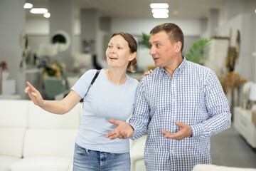 Spouses buyers noticed desired product in furniture store and happily smiles from upcoming purchase. Wife and husband buyer looks around in showroom and chooses right fit furniture