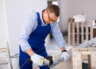 Man in overall and protective glasses using disc saw to cut off metal plank while working in construction site in apartment.