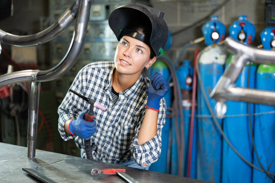 Enthusiastic young female welder in casual plaid shirt, protective gloves and helmet welding metal pipe structure in workshop, holding torch and carefully inspecting quality of weld seams