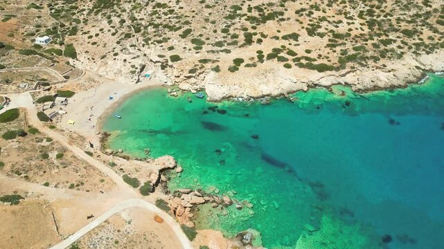 A small, crescent-shaped beach on Donousa Island in the Cyclades, Greece. The beach is surrounded by rocky hills and clear, turquoise water.