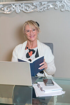 Una mujer rubia sentada en su escritorio, absorta en la lectura de un libro. La escena transmite tranquilidad y concentraci&oacute;n, con la luz natural iluminando suavemente el entorno. 