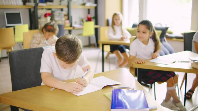 An elementary school boy is doing classwork in his notebook, and his classmate, a girl, is asking him for a pen.