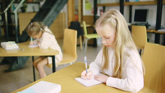 An elementary school girl is doing classwork in her notebook and asking her classmate, another girl, for help.
