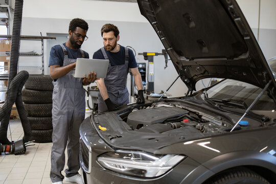 Two automotive mechanics in overalls discussing car repair using digital tablet in auto service workshop. Loft space with tires and tools seen in background.