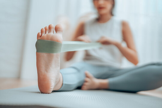Young Asian woman doing pilates workout using elastic strap pulling with arms for shoulder training on yoga mat indoors. Resistance band exercise at home