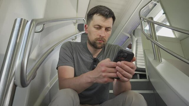 Young man travels on double decker train and sits on flyer using his smartphone. No available seating during railroad trip. Male commutes in dual deck train seated on flyer and surfing internet