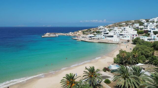 A tranquil, clear-water cove in the Cyclades Islands. The beach is fringed with white sand, lush greenery, and the charming, whitewashed buildings of Donousa.