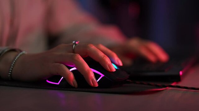 Closeup of unrecognizable female gamer playing video game at computer clicking modern wireless computer mouse in dark virtual room with neon light. Close-up of freelancer woman working late at home.