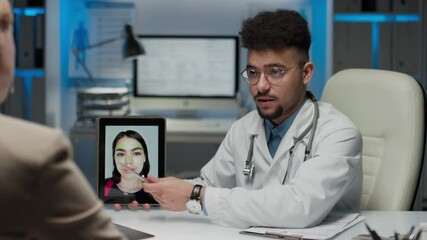Medium pan shot of male Biracial cosmetic surgeon holding tablet with photo of woman and telling unidentified female patient about ways of face contour correction in private clinic - Powered by Adobe