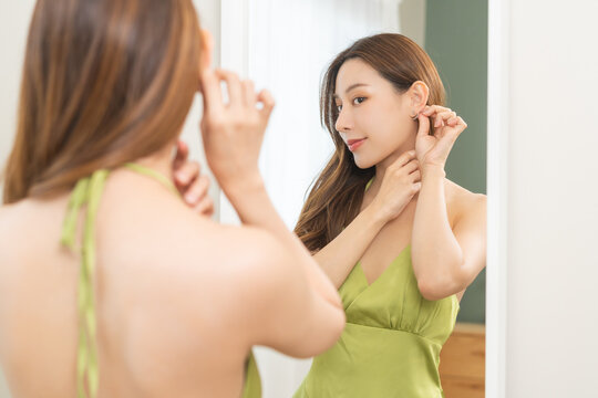 Get dress, elegant asian young woman with makeup glamour, getting ready wearing green cloth, gorgeous female putting earring or jewelry looking into the mirror at home, prepare going event night party