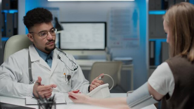 Medium pan shot of young positive bearded physician in glasses and lab coat with stethoscope over neck taking female patients blood pressure using tonometer in modern hospital