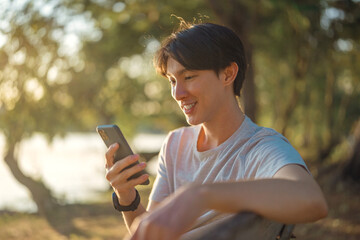 Happy young asian man using smartphone while sitting on bench in public park.